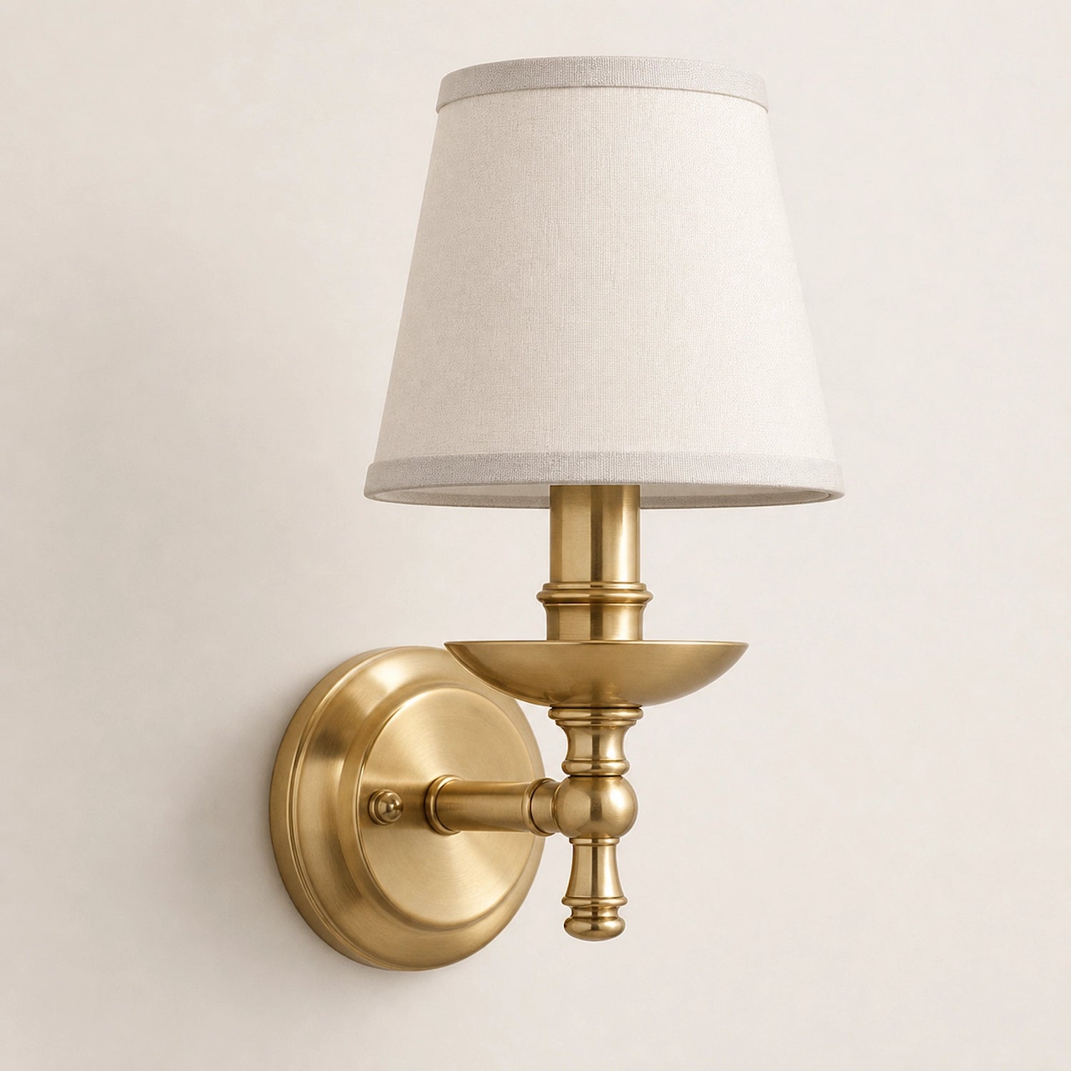 A wall-mounted sconce with a warm brass/gold metal fixture and a tapered off-white fabric shade, shown against a plain light beige wall.