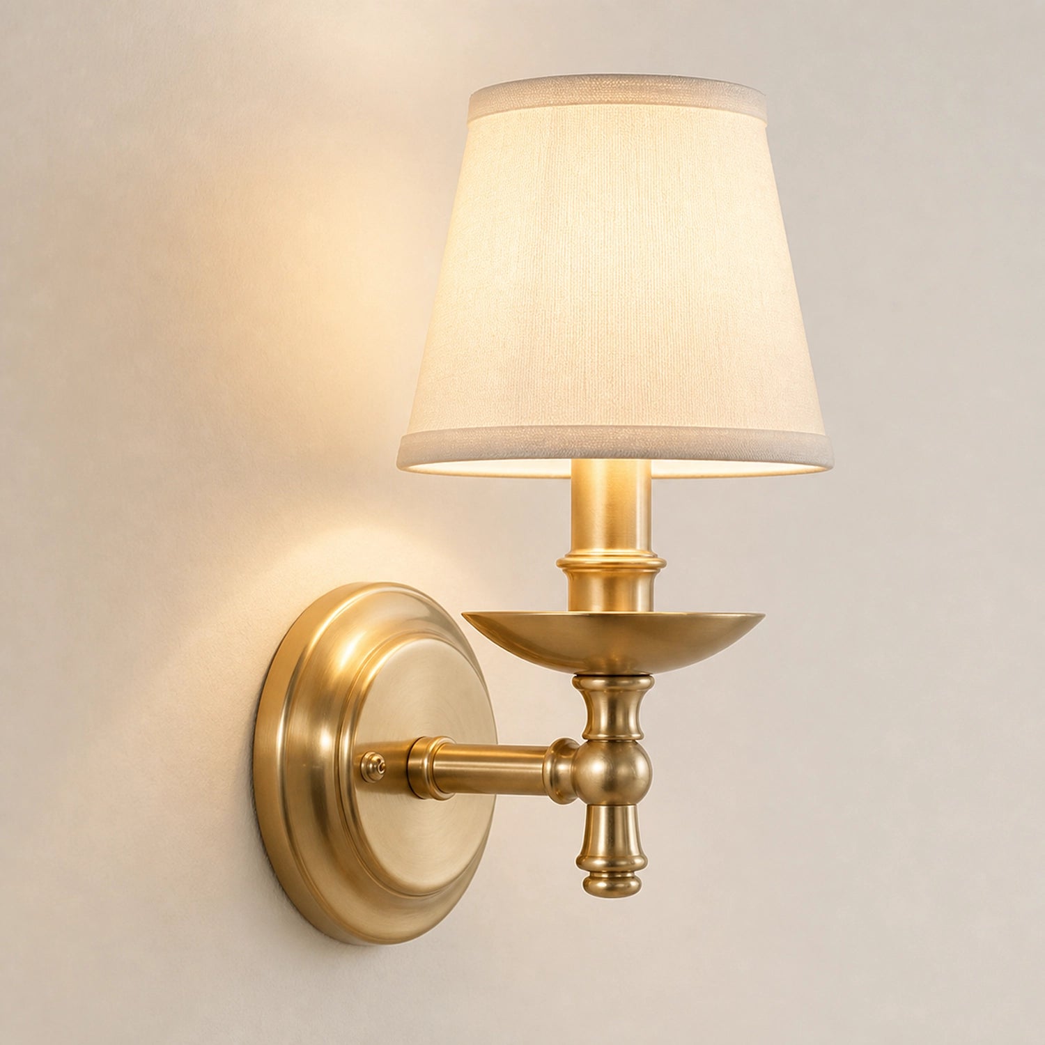 A single wall-mounted sconce with a warm cream fabric drum shade and an antique brass/gold metal fixture, shown lit against a plain light beige wall.