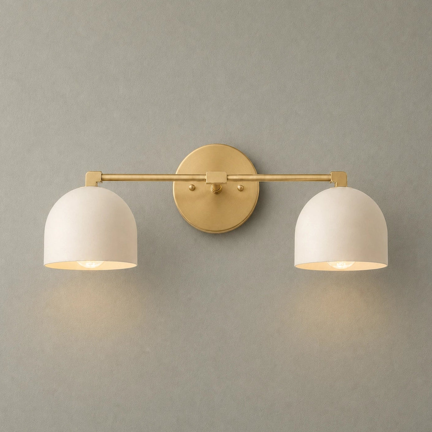A modern two-light wall sconce with a brushed gold metal bar and circular backplate, paired with two white dome-shaped metal shades, shown lit against a textured gray wall.