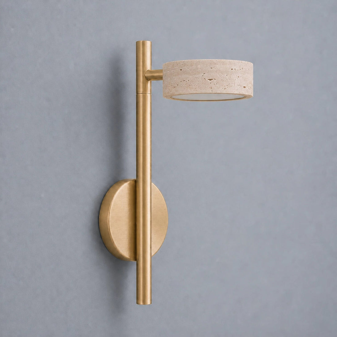 A modern wall-mounted light fixture with a brushed gold metal pole and circular wall plate, paired with a travertine stone shade, shown against a plain gray wall.