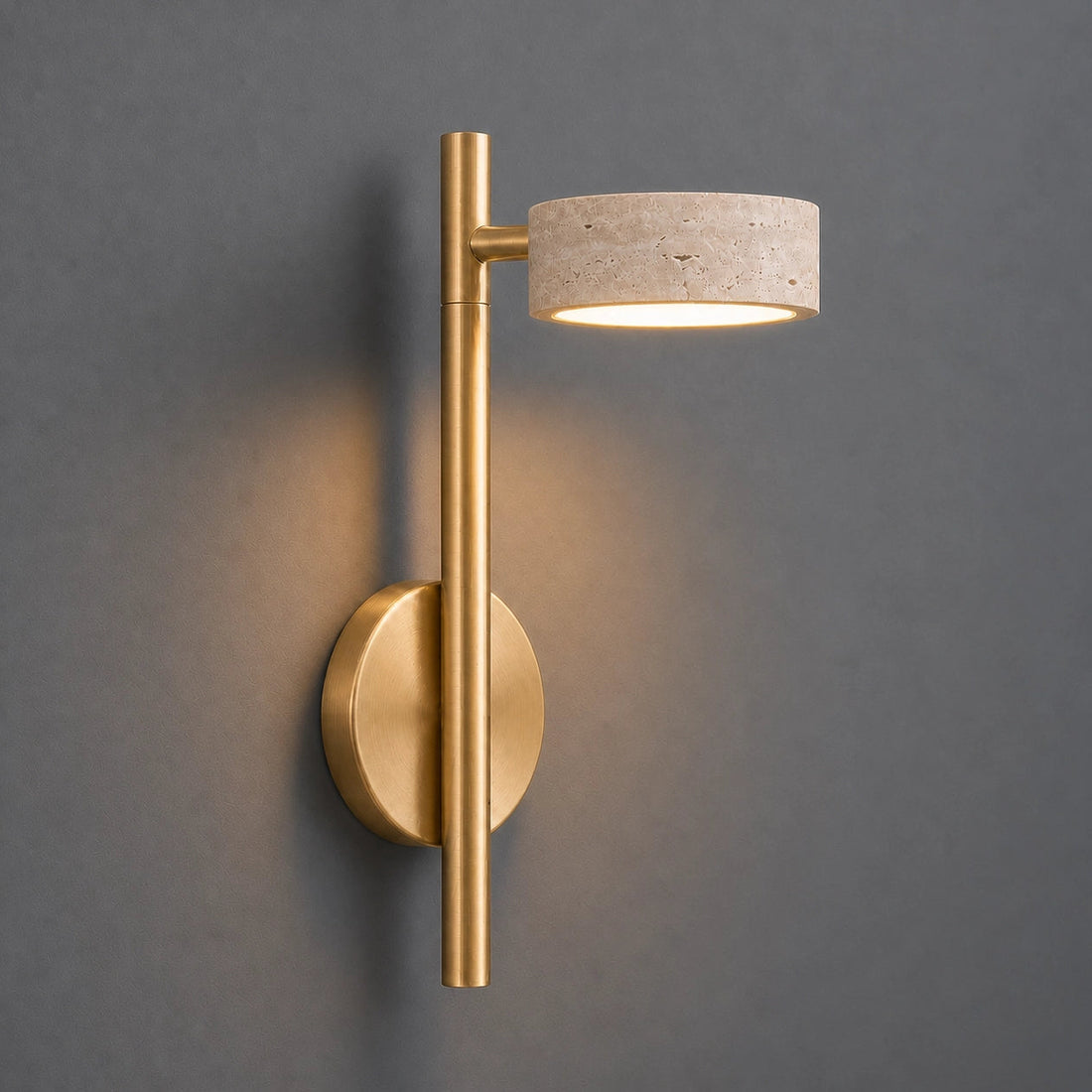 A modern wall-mounted sconce with a brushed gold metal frame and a textured travertine stone shade, shown lit against a dark gray wall.