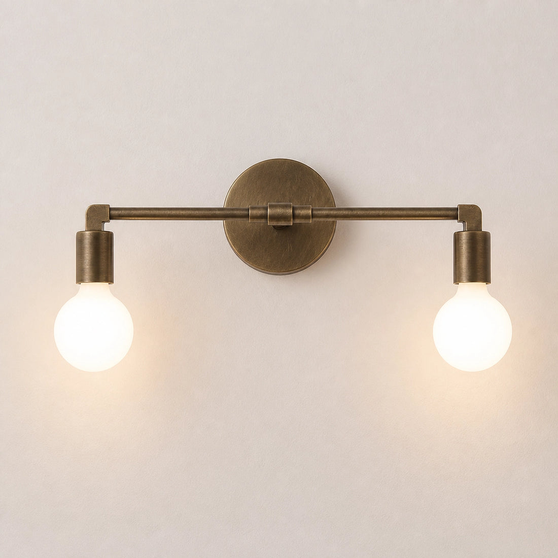 A two-light wall sconce with an antique brass finish and glowing round bulbs, mounted on a plain light beige wall.