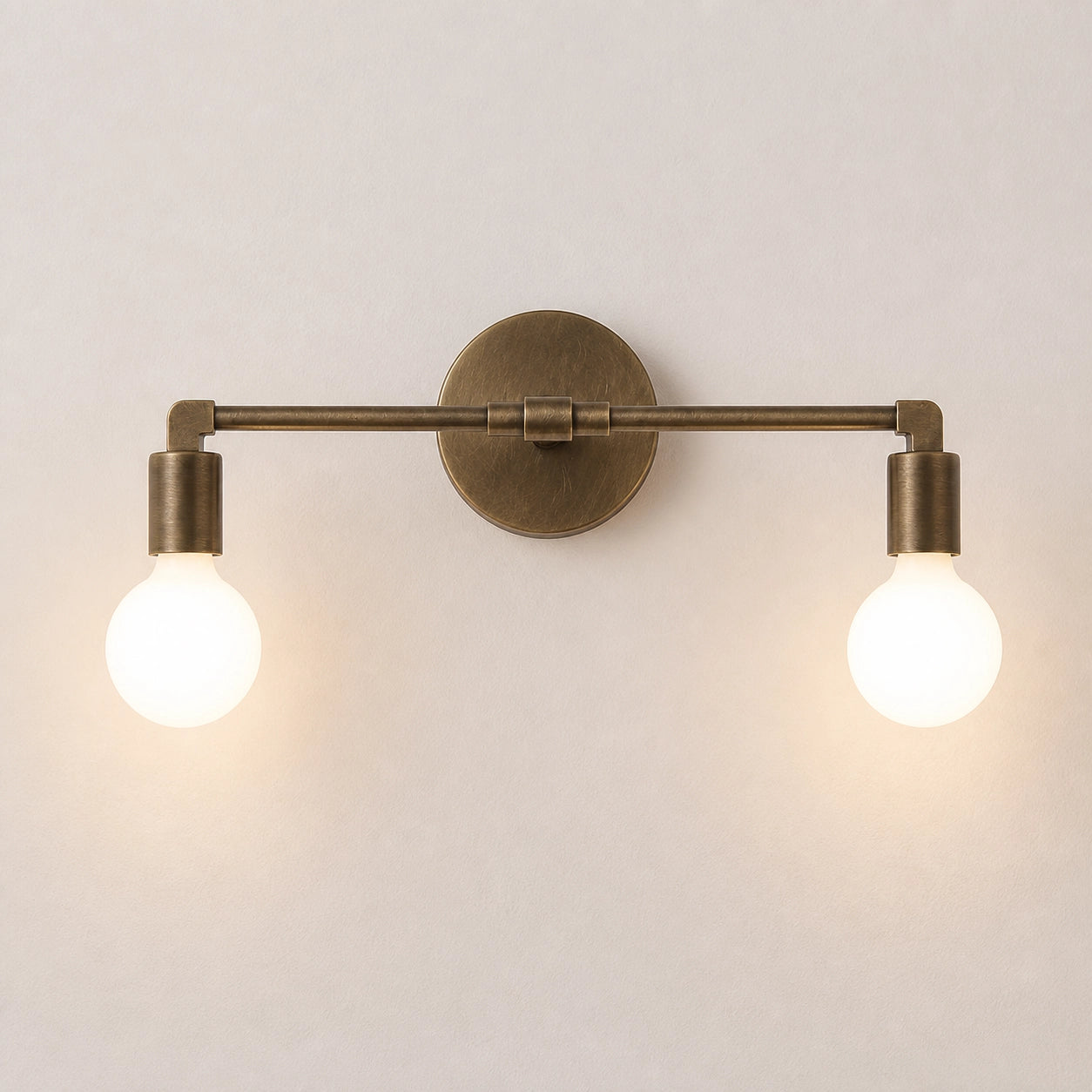 A two-light wall sconce with an antique brass finish and glowing round bulbs, mounted on a plain light beige wall.