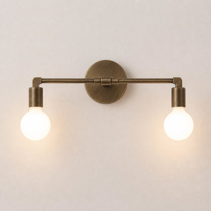 A two-light wall sconce with an antique brass finish and glowing round bulbs, mounted on a plain light beige wall.