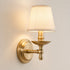 A single wall-mounted sconce with a warm cream fabric drum shade and an antique brass/gold metal fixture, shown lit against a plain light beige wall.