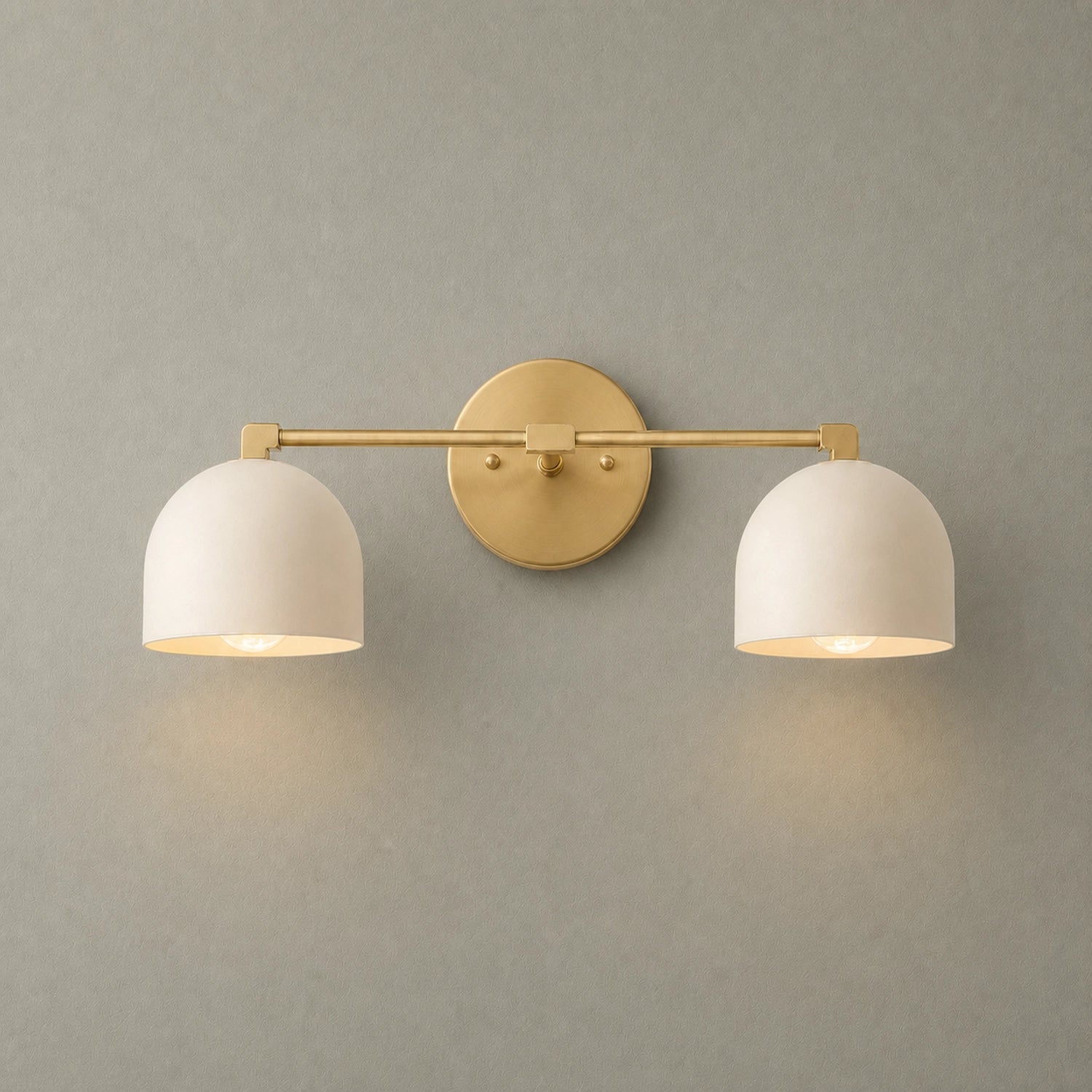 A modern two-light wall sconce with a brushed gold metal bar and circular backplate, paired with two white dome-shaped metal shades, shown lit against a textured gray wall.