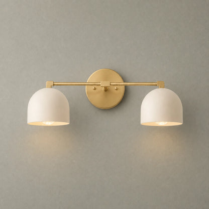 A modern two-light wall sconce with a brushed gold metal bar and circular backplate, paired with two white dome-shaped metal shades, shown lit against a textured gray wall.