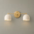 A modern two-light wall sconce with a brushed gold metal bar and circular backplate, paired with two white dome-shaped metal shades, shown lit against a textured gray wall.