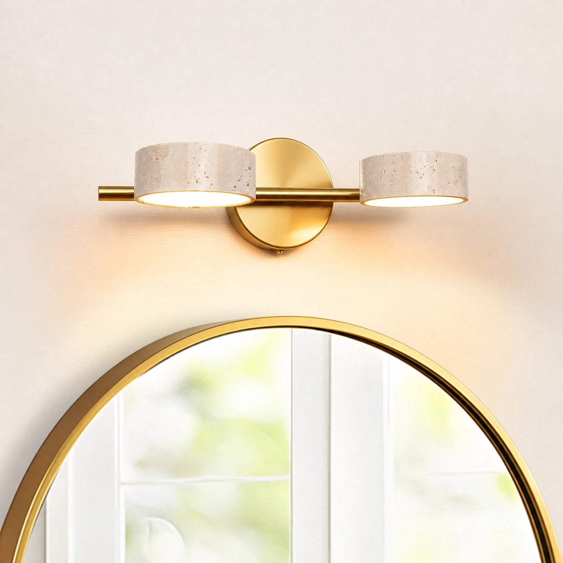 Modern vanity wall light fixture with gold base and two travertine lampshades above a round mirror.