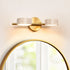 Modern vanity wall light fixture with gold base and two travertine lampshades above a round mirror.